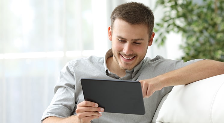 A man relaxes on the sofa looking at a tablet computer and smiling